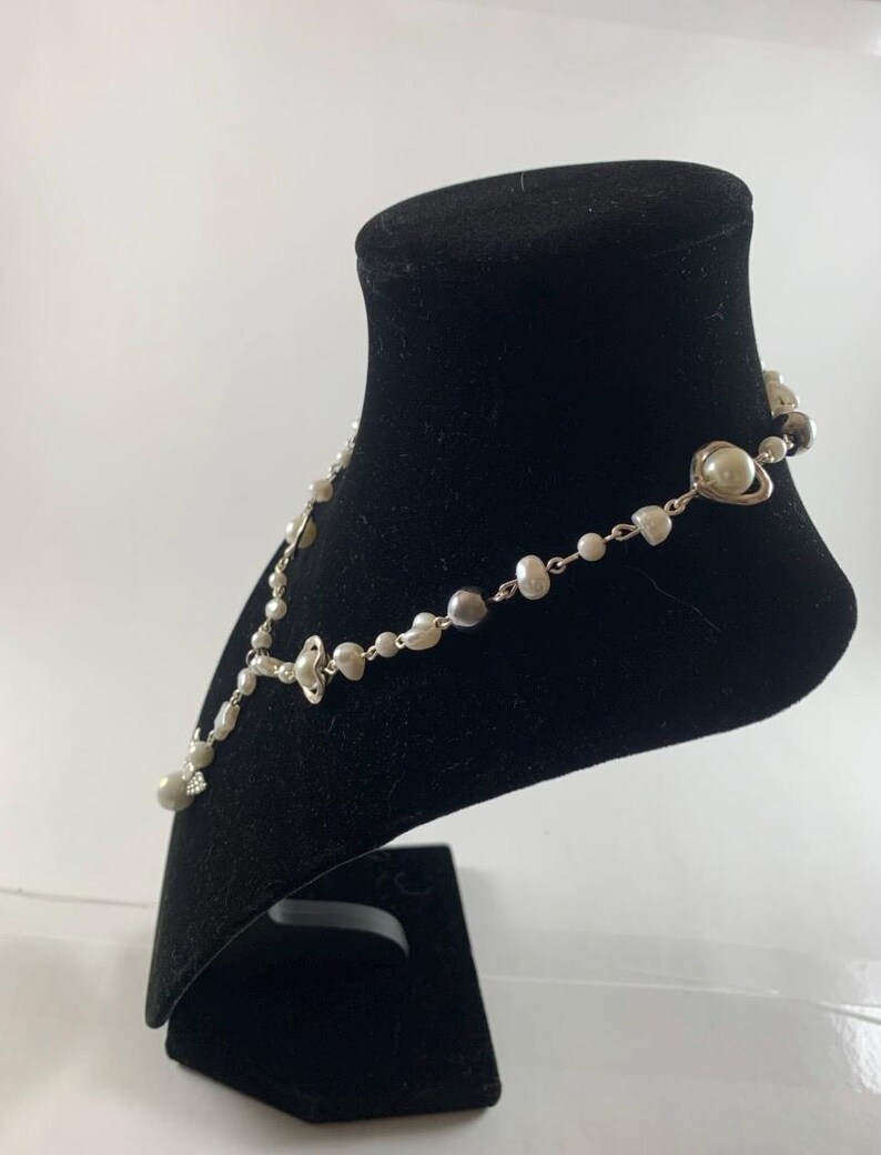 Puede incluir: Un collar de plata con perlas blancas y grises y un colgante en forma de coraz&oacute;n. El collar se muestra en un soporte de terciopelo negro.