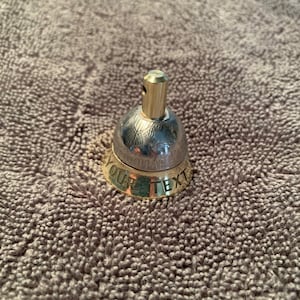 May include: A silver bell with a gold band around the bottom. The band has the words "YOUR TEXT" engraved on it. The top of the bell is silver and has a small hole in the top. The bell is sitting on a brown fuzzy surface.