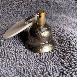 May include: A silver-colored bell-shaped keychain with a gold-colored top and a white tag. The bell is made from a half-dollar coin dated 1971. The keychain is on a gray textured surface.