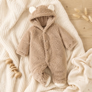 May include: A tan, long-sleeved, hooded one-piece jumpsuit made of fuzzy, plush material. The hood features two small, rounded ears at the top. The garment has a front button closure and white accents on the ear tips and the feet.