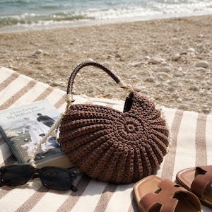 May include: A dark brown crocheted handbag with a shell-like pleated texture, featuring a braided dark brown handle and a decorative white pearl bead strap. The bag is positioned next to a book titled My Brilliant Friend, sunglasses, and sandals.