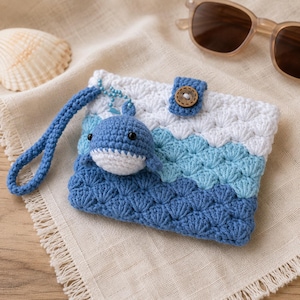 May include: A rectangular crochet pouch featuring a wave-like pattern in white, light blue, and dark blue yarn. The pouch includes a blue crochet wrist strap, a small wooden button closure, and a dangling blue and white crocheted whale charm.
