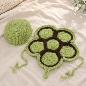 May include: A light green crochet beanie hat paired with a matching flat crochet piece featuring a dark brown and light green hexagonal pattern. The flat piece has six rounded edges and includes two thin crochet ties extending from the sides.