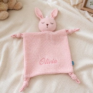 Könnte beinhalten: Eine rosa Baby-Schmusedecke mit einem Hasenkopf und geknoteten Ecken. Der Name "Olivia" ist in passendem Rosa auf die Decke gestickt. Der Hase hat geschlossene Augen und lange Ohren.