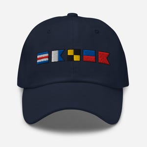 Puede incluir: Gorra de béisbol azul marino con banderas de señales náuticas bordadas en rojo, blanco, azul y amarillo. La gorra tiene una visera curva y una correa ajustable en la parte trasera, ofreciendo un estilo clásico e informal.
