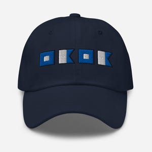 Puede incluir: Gorra de béisbol azul marino con banderas de señales náuticas bordadas en blanco y azul. La gorra tiene una visera curva y una correa ajustable en la parte trasera. El diseño presenta cuatro banderas de señales.