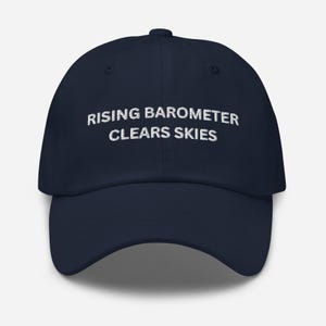May include: A navy blue baseball cap with the text "RISING BAROMETER CLEARS SKIES" in white. The cap has a curved brim and a rounded crown. The hat is made of a soft material.