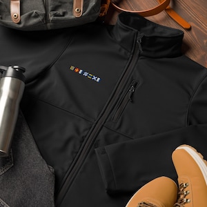 Puede incluir: Una chaqueta softshell negra con cierre de cremallera y un pequeño bolsillo en el lado izquierdo. La chaqueta tiene un diseño de bandera colorido en el pecho izquierdo.