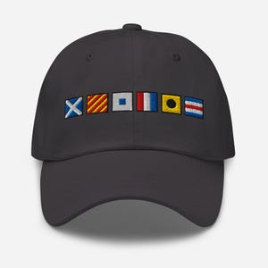 Puede incluir: Gorra de béisbol gris oscuro bordada con seis banderas de señales náuticas de varios colores. Las banderas están dispuestas horizontalmente en la parte delantera. La gorra tiene una visera curva y un botón en la parte superior.