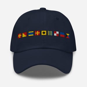Puede incluir: Gorra de béisbol azul marino con banderas de señales náuticas bordadas en varios colores. Las banderas están dispuestas horizontalmente en la parte delantera. La gorra tiene una visera curva y un botón en la parte superior.