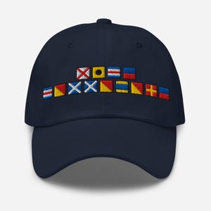 May include: Navy blue baseball cap with embroidered nautical signal flags in various colors. The cap features a curved brim and an adjustable strap for a comfortable fit. A stylish accessory for maritime enthusiasts.