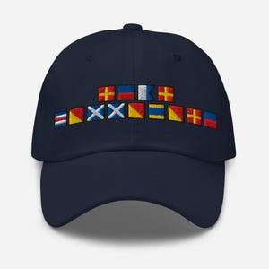 May include: Navy blue baseball cap with embroidered nautical signal flags across the front. The flags are in various colors, including red, yellow, blue, and white. The cap has a curved brim and an adjustable strap.