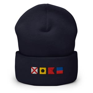 Pode incluir: Gorro azul marinho com bandeiras de sinais náuticos bordadas. As bandeiras são vermelhas, amarelas e azuis, adicionando um toque de cor ao gorro escuro. Um acessório quente para os amantes do mar.