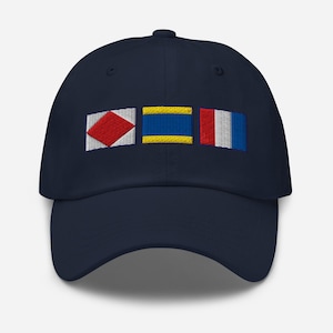 May include: Navy blue baseball cap with embroidered nautical signal flags. The flags feature red, yellow, and blue colors. The cap has a curved brim and a fabric strap closure.