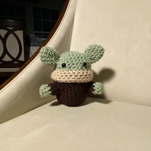 May include: A crocheted green and brown plush toy with a beige face mask. The toy has large ears and is sitting on a white chair.
