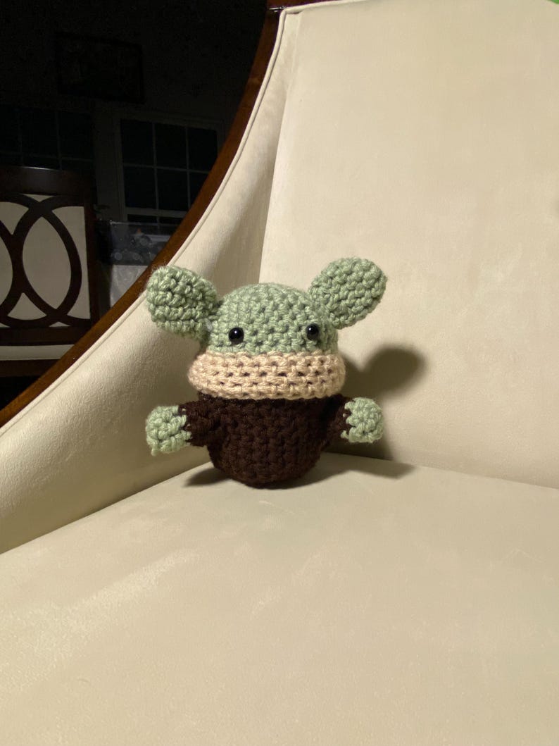 May include: A crocheted green and brown plush toy with a beige face mask. The toy has large ears and is sitting on a white chair.