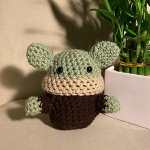 May include: A crocheted green, brown, and beige plush toy resembling a character from the Star Wars franchise. The toy has large ears, black eyes, and a small mouth.