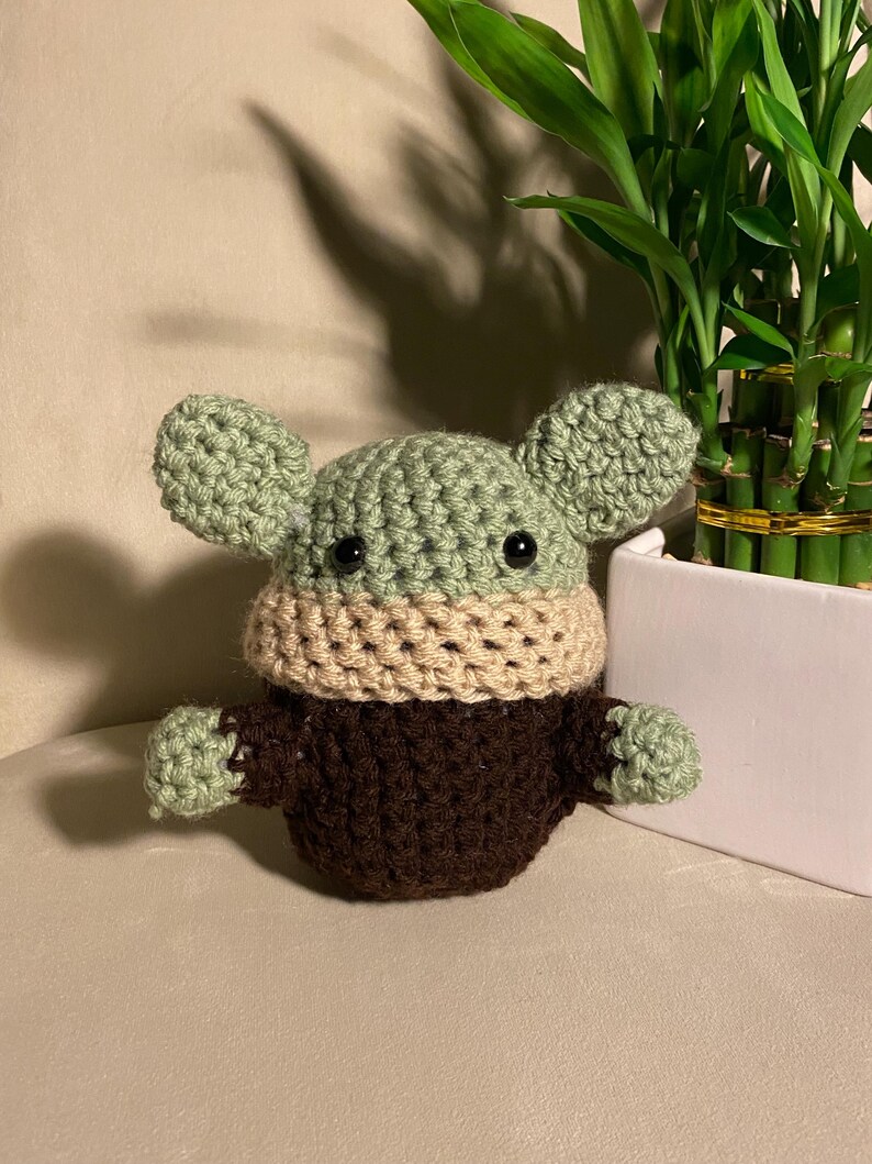 May include: A crocheted green, brown, and beige plush toy resembling a character from the Star Wars franchise. The toy has large ears, black eyes, and a small mouth.