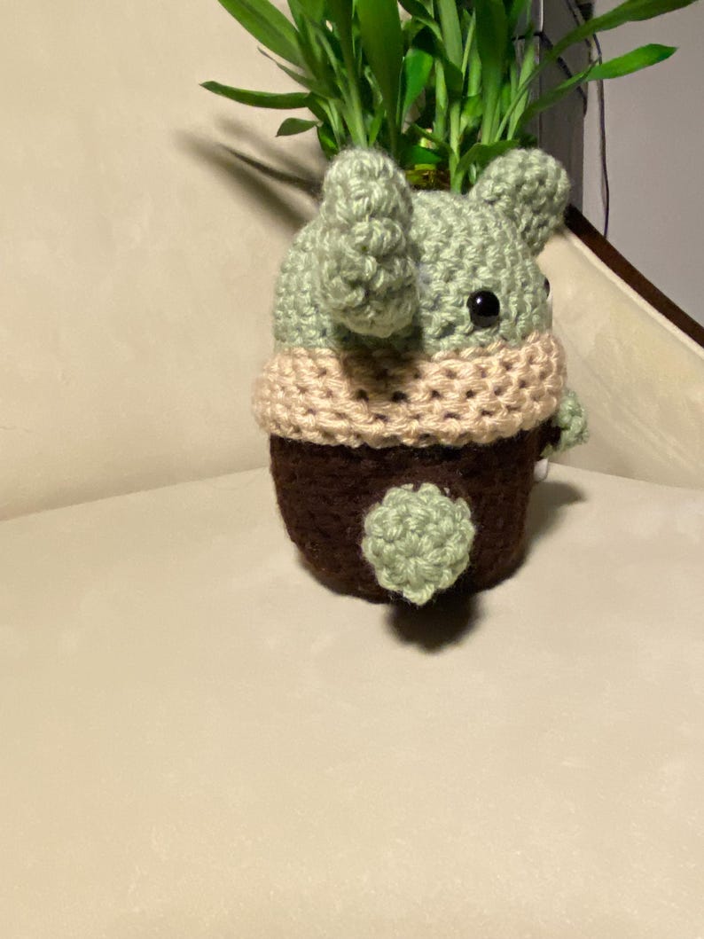 May include: A crocheted green and brown planter shaped like a character from the Star Wars franchise. The planter is decorated with a tan crocheted band around the top.