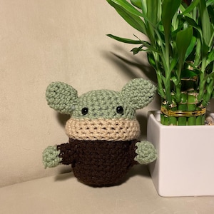 May include: A green crocheted plush toy with brown and beige accents. The toy has large ears and a small mouth. It is sitting on a beige surface next to a potted plant.