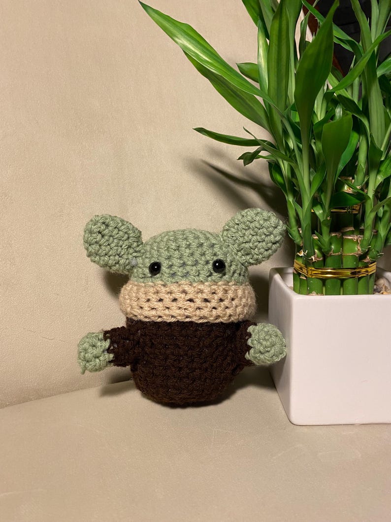 May include: A green crocheted plush toy with brown and beige accents. The toy has large ears and a small mouth. It is sitting on a beige surface next to a potted plant.
