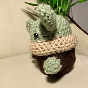May include: A crocheted green and brown plush toy with a tan scarf. The toy is sitting on a light brown surface.