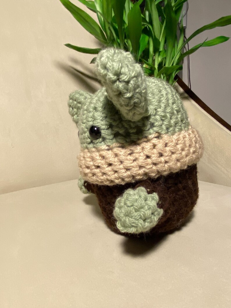 May include: A crocheted green and brown plush toy with a tan scarf. The toy is sitting on a light brown surface.