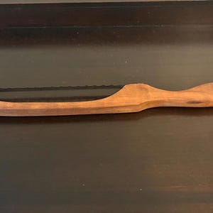 May include: A wooden bread knife featuring a long, curved handle and a thin, serrated metal blade. The handle is crafted from light brown wood with a visible grain pattern, extending along the top and bottom to frame the metal cutting edge.