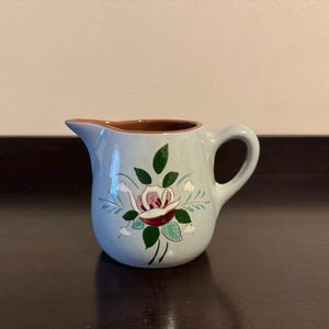 May include: A small, light blue ceramic pitcher with a handle and spout. The pitcher is decorated with a floral design featuring a pink and red rose with green leaves. The interior rim is brown.