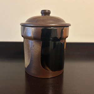 May include: A ceramic jar with a lid, featuring a brown and black color scheme. The jar has a cylindrical shape with a lid and a small knob. The exterior has a textured, glazed finish with dark brown and black vertical patterns.