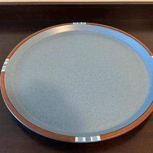 May include: A round, speckled blue ceramic plate with a brown rim. The rim features a decorative band of blue, white, and brown stripes. This plate is suitable for serving food and is a decorative kitchen item.