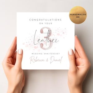 May include: A white card with the text "Congratulations on your 3 Leather Wedding Anniversary" and the names "Rebecca & Daniel" in a script font. The card has a watercolor-style design with pink accents and is held by two hands.