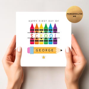 May include: A white card with the text "HAPPY FIRST DAY OF SCHOOL" above a row of colourful crayons. Below, the word "SCHOOL" is written in rainbow colours, and a yellow pencil with the name "GEORGE" is featured. A gold circle says "PERSONALISE ME".