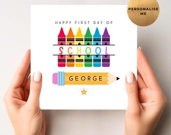 Puede incluir: Una tarjeta blanca con el texto "HAPPY FIRST DAY OF SCHOOL" encima de una fila de crayones coloridos. Debajo, la palabra "SCHOOL" está escrita en colores del arcoíris, y un lápiz amarillo con el nombre "GEORGE" está presente. Un círculo dorado dice "PERSONALISE ME".