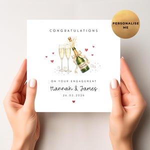 May include: A white engagement card with the words "Congratulations" and "On Your Engagement" printed in black. The card features a watercolour illustration of champagne glasses, a bottle, and hearts. The names "Hannah & James" and the date "26.03.2026" are also printed.