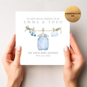May include: A square baby shower card held by hands. The card features a watercolor illustration of baby clothes hanging on a clothesline. Text reads "TO VERY SPECIAL PARENTS TO BE EMMA & THEO ON YOUR BABY SHOWER 19TH JULY 2025". A gold circle says "PERSONALISE ME".