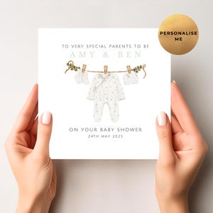 May include: A baby shower greeting card held by hands. The card features a watercolor illustration of baby clothes hanging on a clothesline. Text reads "TO VERY SPECIAL PARENTS TO BE AMY & BEN ON YOUR BABY SHOWER 24TH MAY 2025".