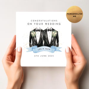 Puede incluir: Una tarjeta de boda con el texto "Felicidades en tu boda". La tarjeta presenta ilustraciones de dos chaquetas de esmoquin con pajaritas y detalles florales. Una cinta azul claro muestra los nombres "Lee & Jack" y la fecha "6 de junio de 2025".