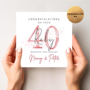 May include: A white card with the text "CONGRATULATIONS ON YOUR 40 Ruby WEDDING ANNIVERSARY Nancy & Peter." The number 40 and the word "Ruby" are in a watercolour style, with a pink hue. A gold circle with the text "PERSONALISE ME" is in the upper right corner.