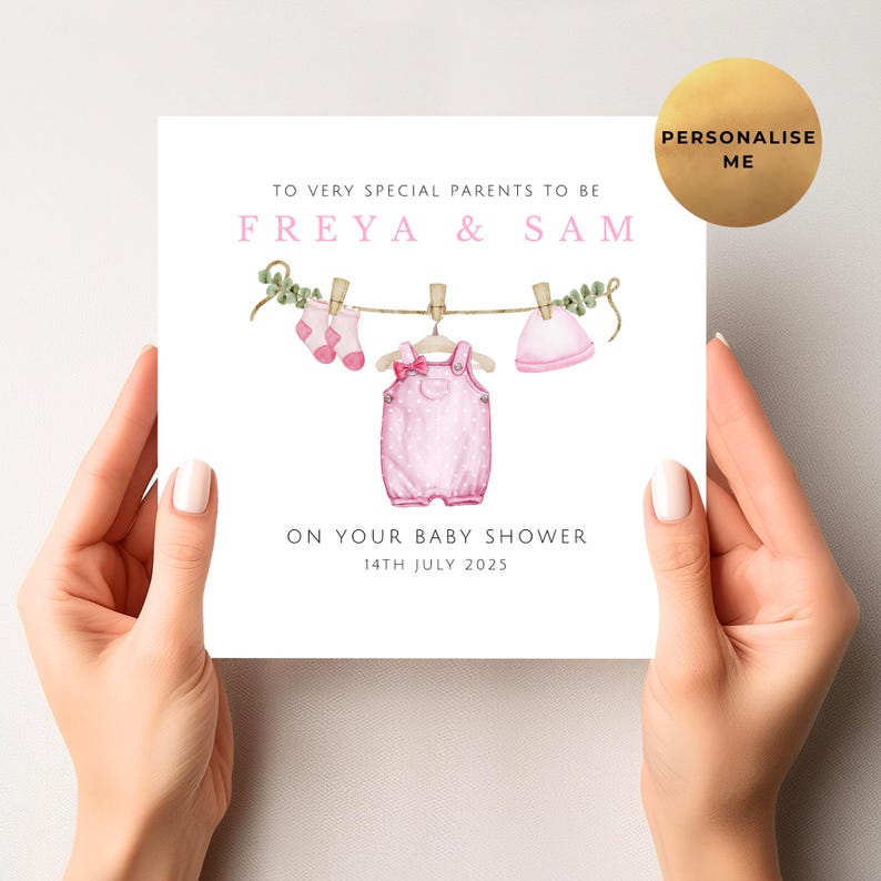 May include: A baby shower card held by hands, featuring a watercolor illustration of baby clothes hanging on a clothesline. The card reads "TO VERY SPECIAL PARENTS TO BE FREYA & SAM" and "ON YOUR BABY SHOWER 14TH JULY 2025". A gold circle says "PERSONALISE ME".