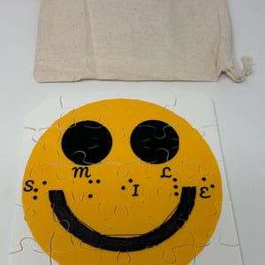 May include: A square jigsaw puzzle featuring a bright yellow circular smiley face with black eyes and a black curved mouth. The word "SMILE" is printed in black letters across the centre of the face. A beige drawstring fabric storage bag is included.