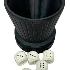 May include: A black dice cup with a textured surface and a set of six ivory-colored dice. Each die has black dots indicating the numbers one through six. The dice are scattered near the cup.