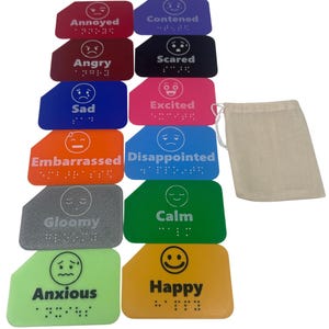 May include: A set of colorful emotion cards with braille text and corresponding facial expressions. The cards are in various colors, including red, purple, and blue, with words like "Angry" and "Happy". A beige drawstring bag is on the right.