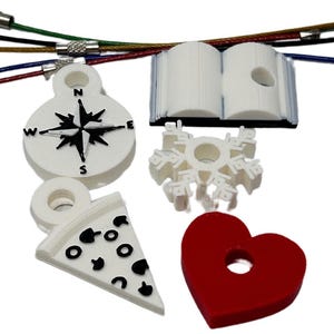 May include: Five white 3D printed charms with a hole for a keychain. The charms are a compass, a snowflake, a book, a pizza slice, and a red heart.