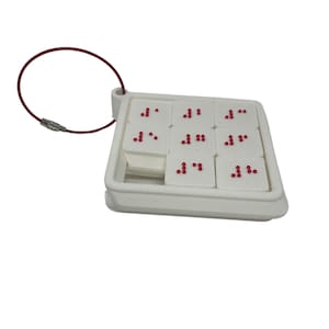 May include: White plastic keychain with a square frame containing nine small white tiles with red dots arranged in Braille patterns.