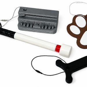 May include: A set of mobility and accessibility tools including a white and black cane with a red band, a gray rectangular block with the text Blind is Mind, a brown and white paw print shaped tag, and a black T-shaped handle with a cord.