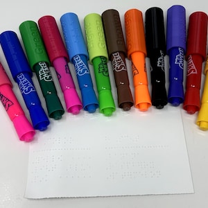 以下が含まれることがあります： ピンク、青、緑、オレンジなど、さまざまな色のカラフルなMr. Sketchマーカーのコレクション。各マーカーにはお揃いのキャップが付いており、ブランド名が見えます。点字の白い紙が前景にあります。