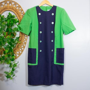 May include: A short-sleeved dress featuring a vibrant green body with a contrasting dark navy blue central panel and matching navy blue patch pockets. The dress is accented with two vertical rows of light-colored buttons down the front center.