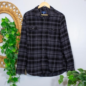 May include: A long-sleeve button-down shirt featuring a black and dark gray plaid pattern. The shirt includes a standard collar, a front button placket, and two chest pockets with buttoned flaps. The fabric has a soft, brushed texture throughout.