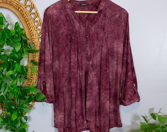 Pode incluir: Uma blusa em tom bordô profundo com um padrão subtil de espirais, decote em V com carcela de botões parcial e mangas três quartos com presilhas abotoadas. A peça tem um corte descontraído com pregas verticais na parte da frente.